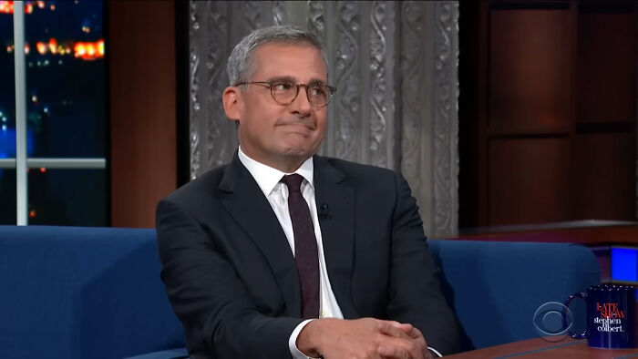 Actor Steve Carell in a suit and glasses, smiling during a heartwarming celebrity encounter on a late-night talk show.
