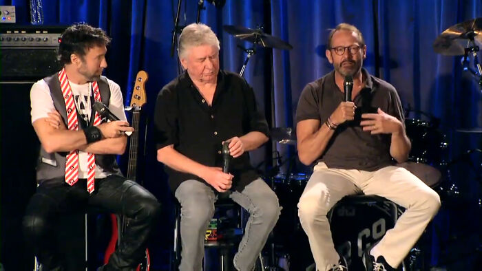 Three men sharing heartwarming celebrity encounters on stage during a live music event with instruments in the background.