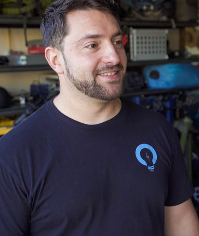 Man with beard smiling, wearing a black shirt with a blue lightbulb logo, pictured in a workshop setting. Man with beard smiling, wearing a black shirt with a blue lightbulb logo, pictured in a workshop setting.