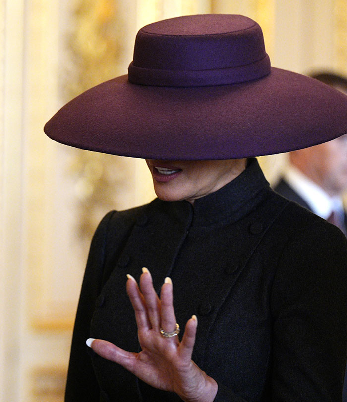 Melania wearing a large purple hat and black outfit during a UK event, outfit widely criticized and mocked. Melania wearing a large purple hat and black outfit during a UK event, outfit widely criticized and mocked.