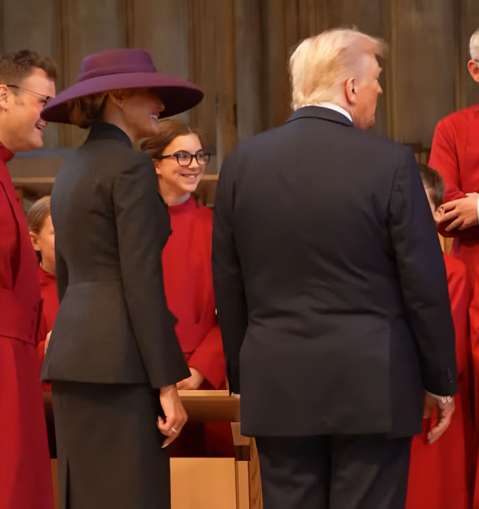 Melania Trump in a dark outfit and large hat standing next to Donald Trump during a UK event with a choir in red robes. Melania Trump in a dark outfit and large hat standing next to Donald Trump during a UK event with a choir in red robes.