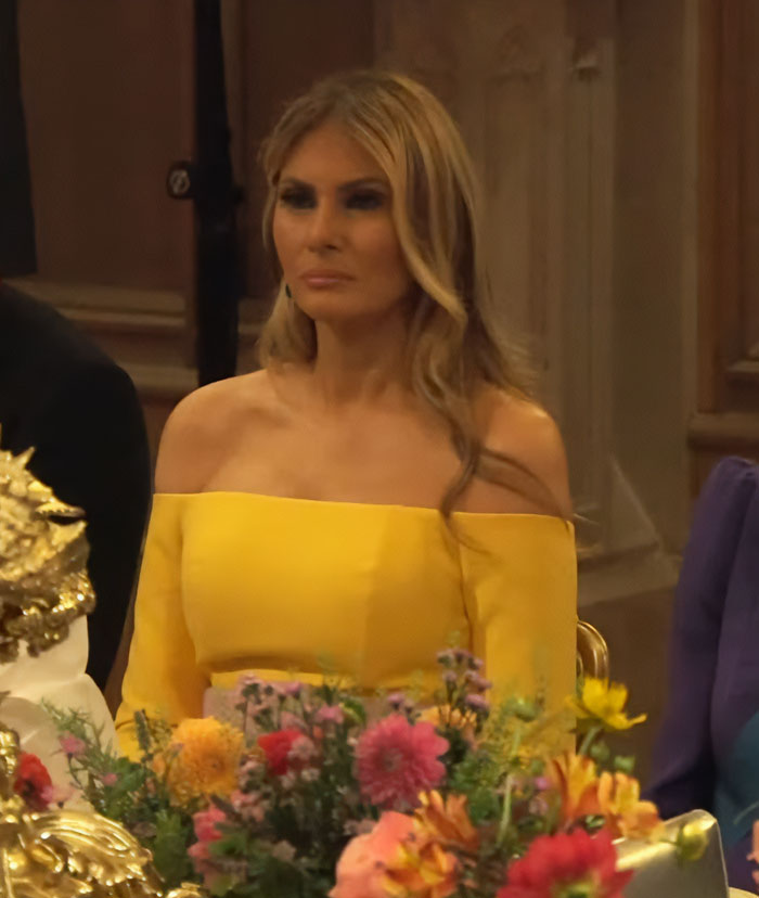 Melania Trump in a yellow off-shoulder dress at a formal UK event with floral decorations in the foreground. Melania Trump in a yellow off-shoulder dress at a formal UK event with floral decorations in the foreground.