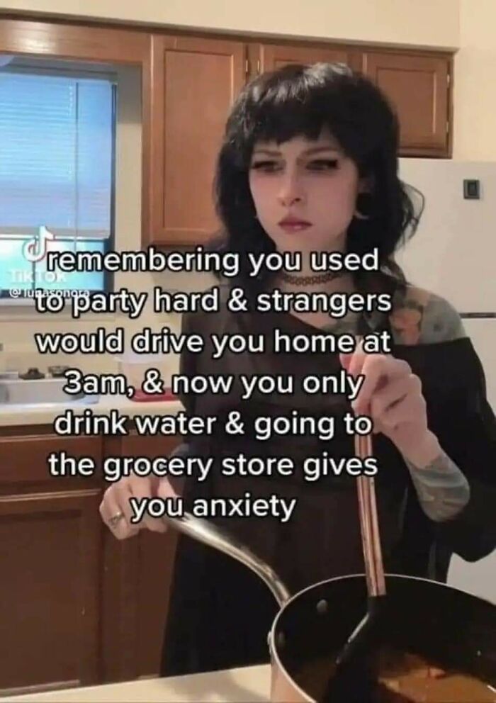 Young person with dark hair and tattoos cooking in a kitchen with a funny mental health meme about anxiety and everyday struggles.