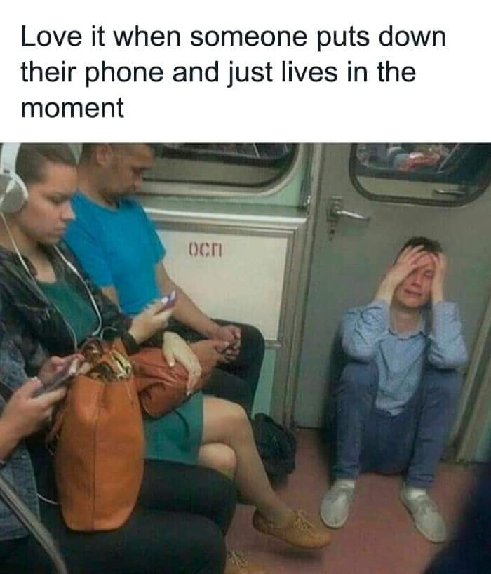Commuters mostly on their phones while one person sits on the floor holding their head, reflecting mental health memes.