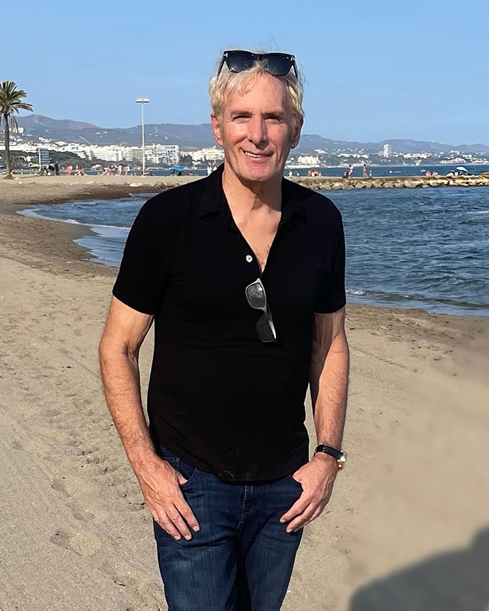 Michael Bolton standing on a beach wearing a black shirt and sunglasses with a scenic ocean background.