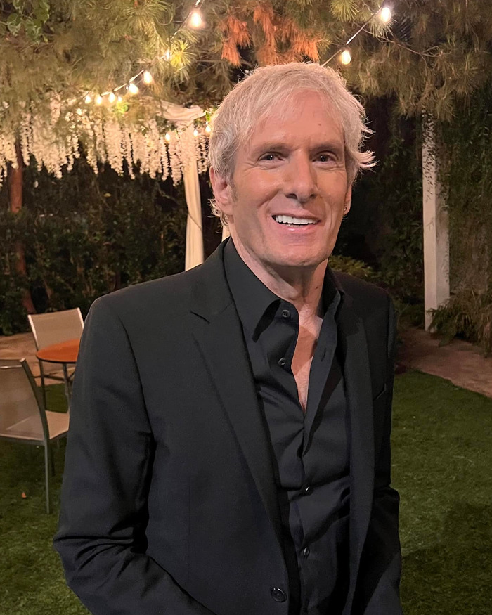 Michael Bolton smiling outdoors at night, wearing a black suit, highlighting a weird symptom before his heartbreaking diagnosis.
