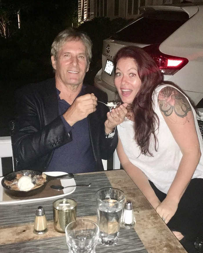 Michael Bolton smiling with a woman at a table, sharing a dessert, highlighting symptom before heartbreaking diagnosis.
