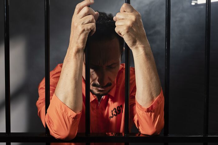 Man in an orange prison uniform holding black metal bars with a somber expression, depicting bizarre sewer worker finds.
