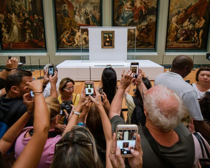 Crowd of people taking photos of art in a museum, illustrating popular travel destinations seen in person versus pictures.