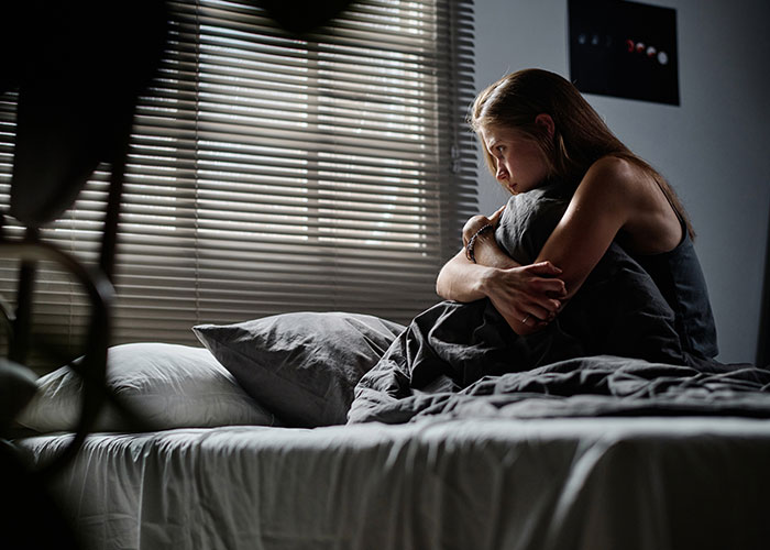 Woman sitting on bed hugging pillow, upset and stressed, highlighting conflict with MIL over triggering allergies. Woman sitting on bed hugging pillow, upset and stressed, highlighting conflict with MIL over triggering allergies.