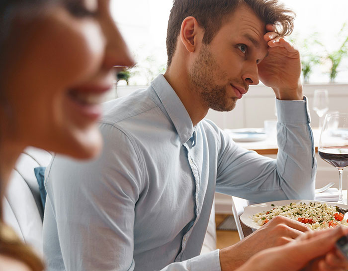 Young man in a light blue shirt looking uncomfortable at the dining table with food and a woman smiling nearby.