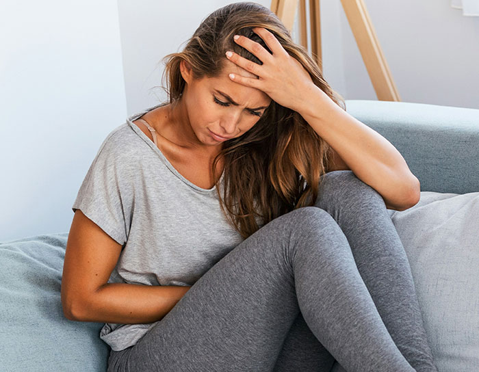 Woman in casual clothes sitting on couch, holding stomach and head, showing discomfort from spoiled food fed by cheap MIL.