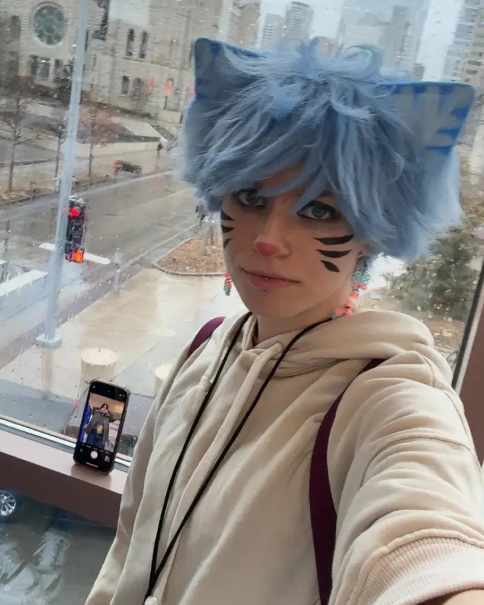 Person dressed as a furry with blue cat ears and face paint taking a selfie by a large window in an urban setting.