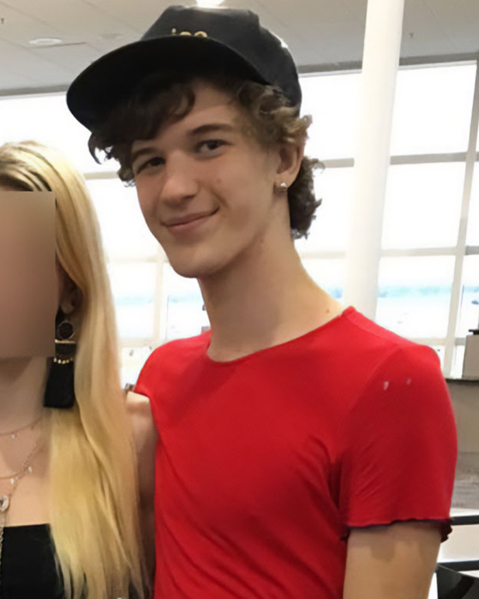 Young man in red shirt and black cap standing indoors next to a woman with blurred face, connected to Minneapolis suspect story.