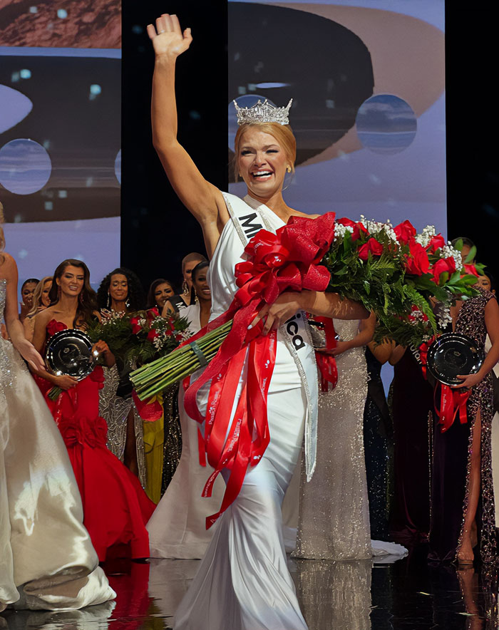Miss America winner wearing crown and sash, holding bouquet, waving to crowd amid pageant participants on stage.