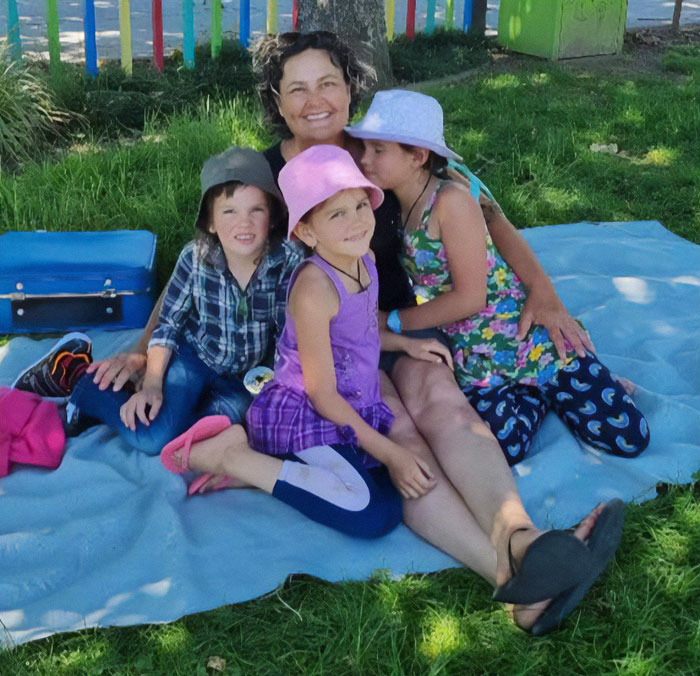 Mother sitting on a blanket outdoors with three children, related to fugitive dad who vanished into wilderness with kids.