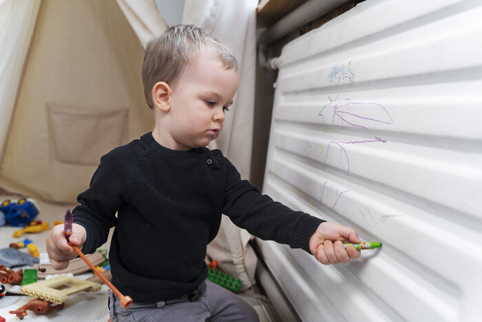 Toddler boy drawing on wall with crayons, showing signs of being out of control after joining nursery, concerned mom nearby. Toddler boy drawing on wall with crayons, showing signs of being out of control after joining nursery, concerned mom nearby.