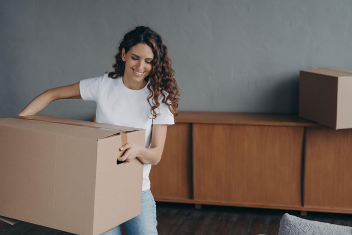 Teen carrying cardboard box inside room, smiling and organizing during a move or unpacking process.