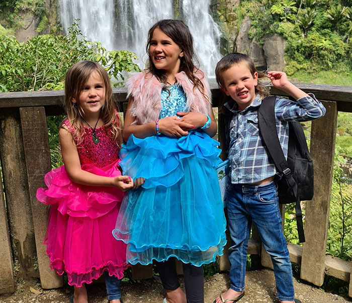 Three missing kids dressed in bright clothes standing by a wooden railing with a waterfall in the background.