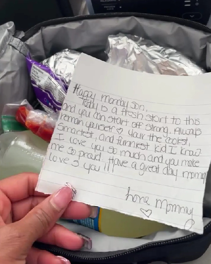 Hand holding a loving handwritten note inside a child's lunchbox with wrapped food and snacks visible.