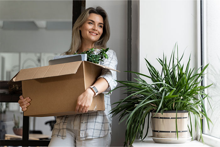 Smiling woman carrying a box of office items, depicting a quitting moment with a golden reason behind it. Smiling woman carrying a box of office items, depicting a quitting moment with a golden reason behind it.