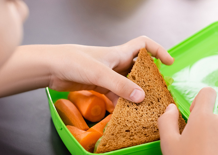 Child holding sandwich near cut carrots in green lunchbox representing mom pulling son from school after snack ban.