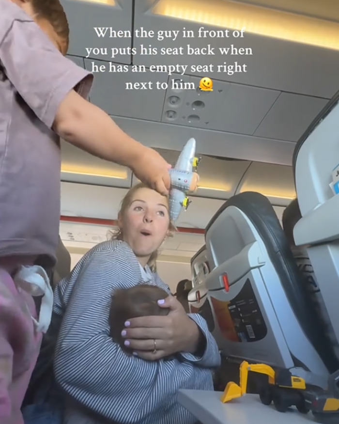 Mom looks annoyed holding baby on plane with reclined seat pressing against her, sparking viral debate about seat space. Mom looks annoyed holding baby on plane with reclined seat pressing against her, sparking viral debate about seat space.
