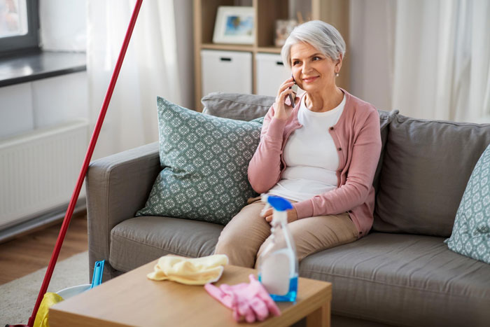 Older woman sitting on a couch talking on the phone, with cleaning supplies nearby, reflecting a daughter bolts up room scene.