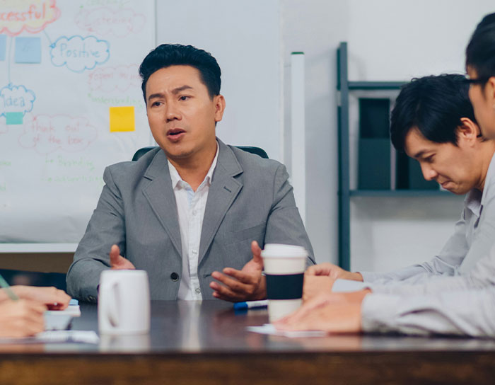 Man in gray suit talking during a business meeting with colleagues, discussing TikTok moms and professions disliked.