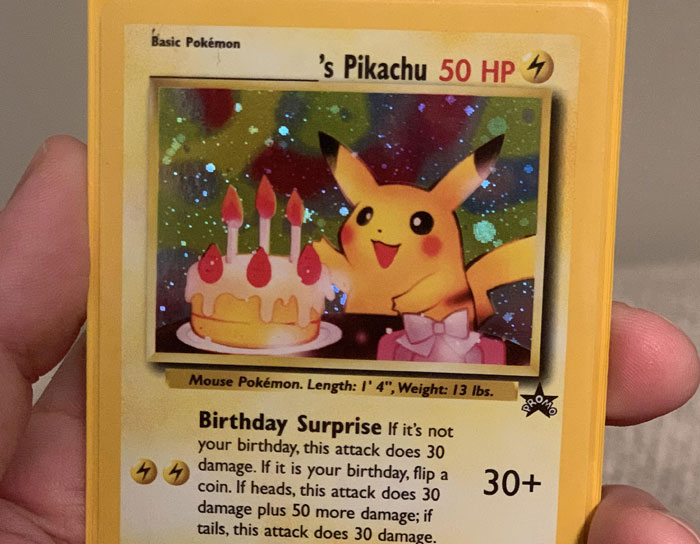 Hand holding a holographic Pikachu Pokémon card with 50 HP and birthday cake illustration.