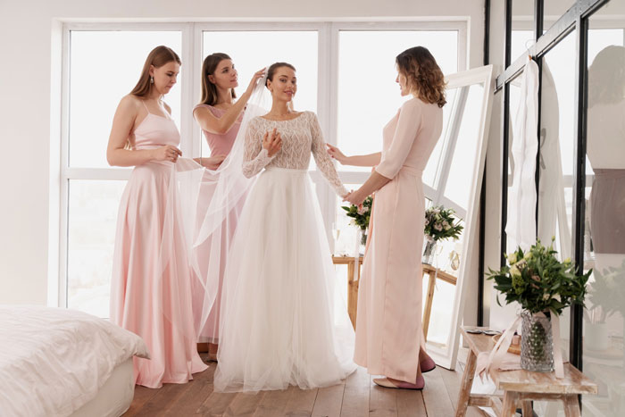 Bride in white dress with bridesmaids in pink gowns preparing for wedding in a bright room with flowers and mirrors. Bride in white dress with bridesmaids in pink gowns preparing for wedding in a bright room with flowers and mirrors.