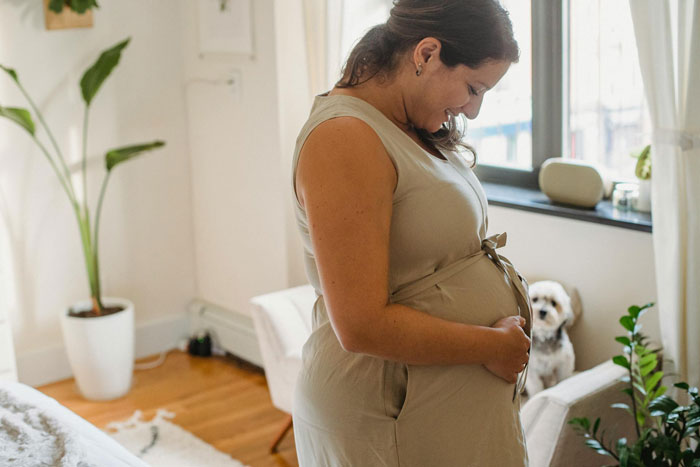 Pregnant woman in casual dress smiling and holding belly at home, highlighting adult kids expected to help out.