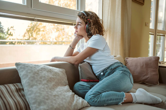 Young woman wearing headphones sitting on couch looking out window, reflecting on siblings fight over house they share.