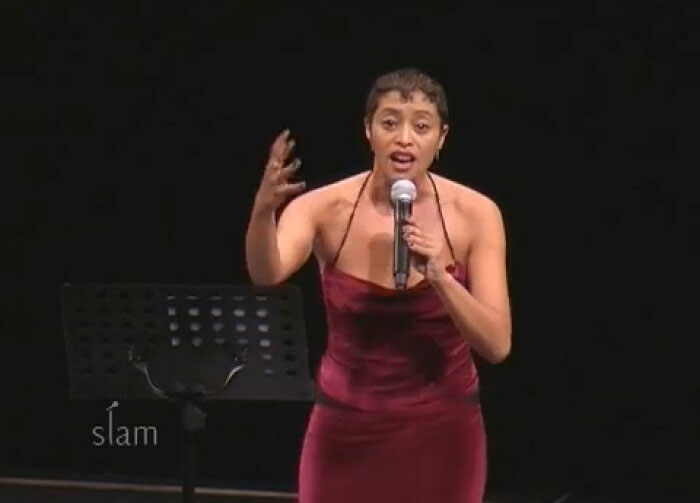 Female performer in a red dress speaking passionately into a microphone on stage at a slam event related to celebrities who vanished.