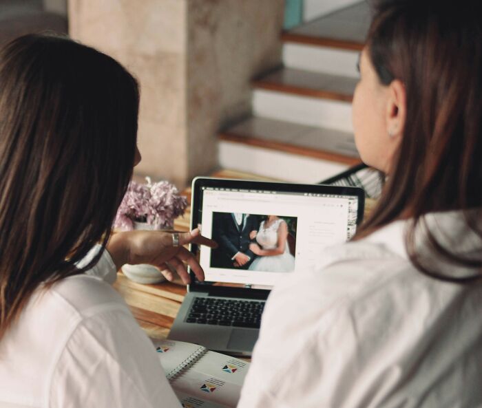 Two women reviewing wedding photos on a laptop, discussing professions that raise suspicion when combined.