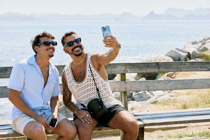 Two men sitting on a bench by the sea, smiling and taking a selfie, reflecting on school bullies and their lives.