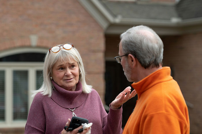 Woman holding phone talking to man outside house, illustrating neighbor wars with Greek chanting music dispute. Woman holding phone talking to man outside house, illustrating neighbor wars with Greek chanting music dispute.