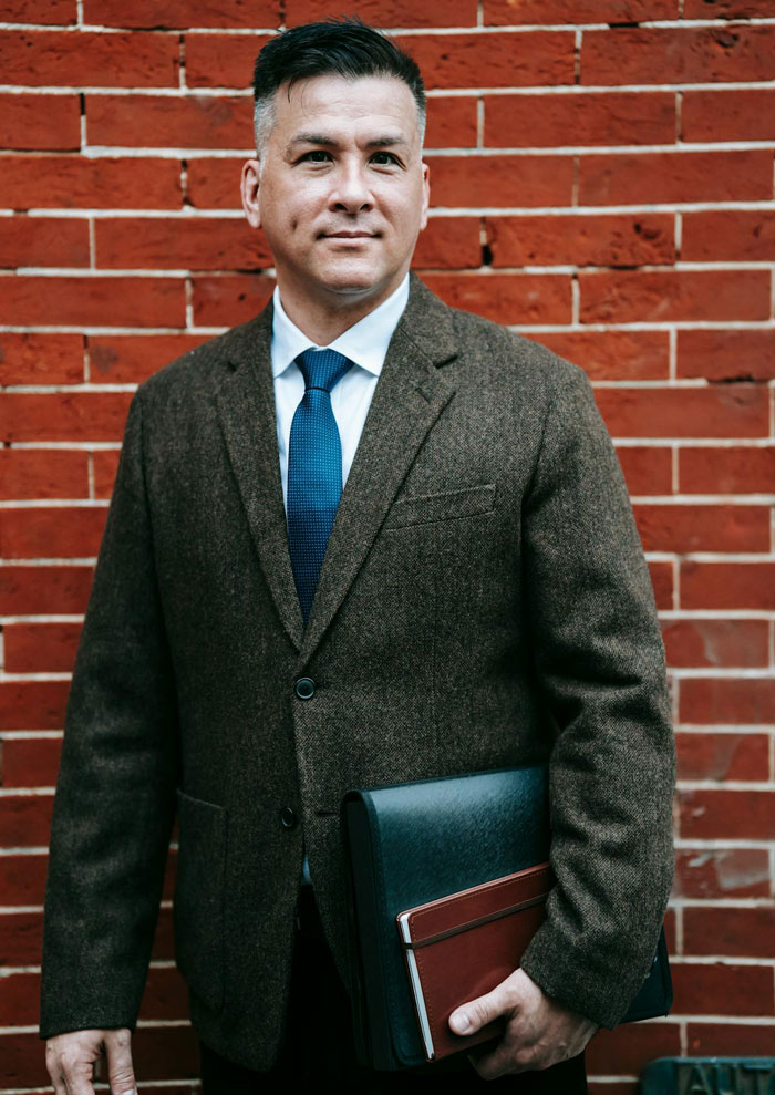 Man in brown blazer and blue tie standing against a brick wall holding a folder, related to neighbor wars and Greek chanting music. Man in brown blazer and blue tie standing against a brick wall holding a folder, related to neighbor wars and Greek chanting music.