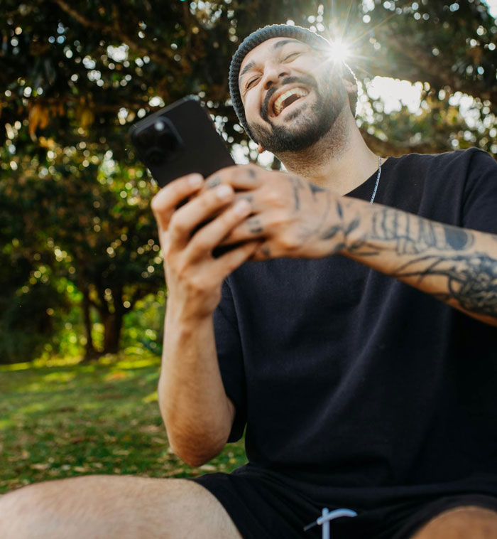 Man with tattoos and beanie laughing outdoors, holding phone while blasting 1900s Greek chanting music in neighbor wars. Man with tattoos and beanie laughing outdoors, holding phone while blasting 1900s Greek chanting music in neighbor wars.