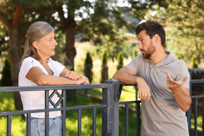 Neighbor upset talking with man over nanny parking issue in front of home near a metal gate on a sunny day