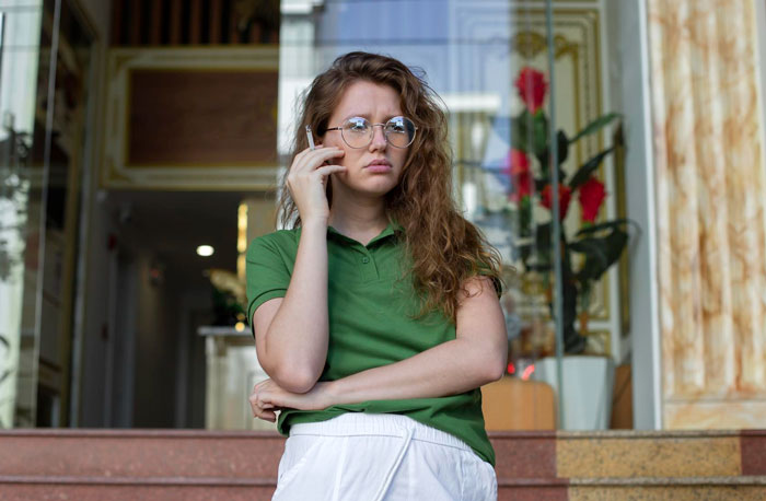 Young woman upset on phone standing outside home, representing neighbor upset over nanny parking in front of house