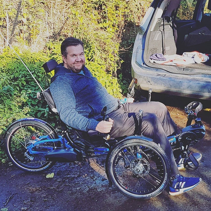 Man with prosthetic legs smiling while sitting on a recumbent tricycle near a parked car outdoors. Man with prosthetic legs smiling while sitting on a recumbent tricycle near a parked car outdoors.