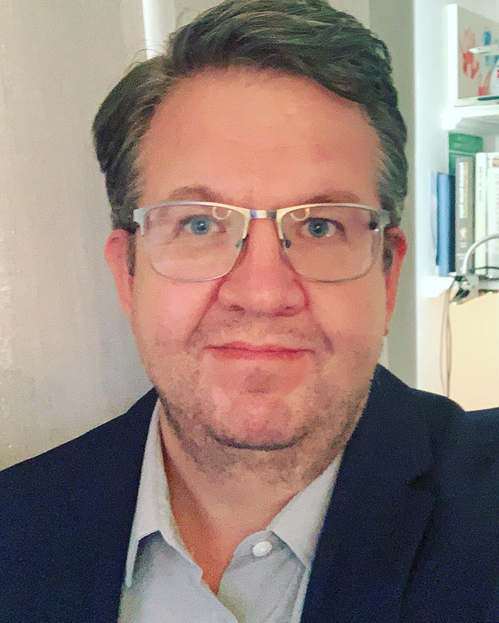 Man wearing glasses and a blue shirt in an indoor setting, representing surgeon who intentionally had own legs amputated. Man wearing glasses and a blue shirt in an indoor setting, representing surgeon who intentionally had own legs amputated.