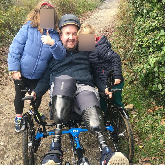 Man with dual leg amputations sitting outdoors on a recumbent bike with two children, smiling and enjoying a walk. Man with dual leg amputations sitting outdoors on a recumbent bike with two children, smiling and enjoying a walk.