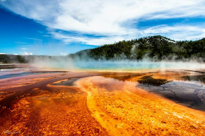 Colorful hot spring at a travel destination with steam rising and a forested hill under a partly cloudy sky.