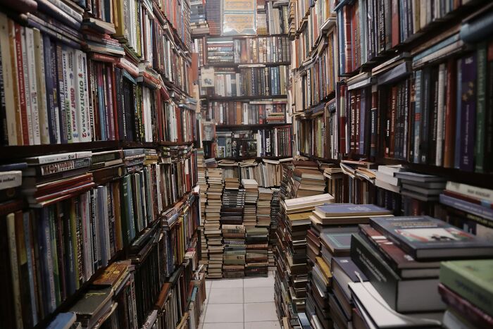 Narrow bookstore aisle filled with stacked books on shelves and floor, evoking stories from people who explore beneath our feet.