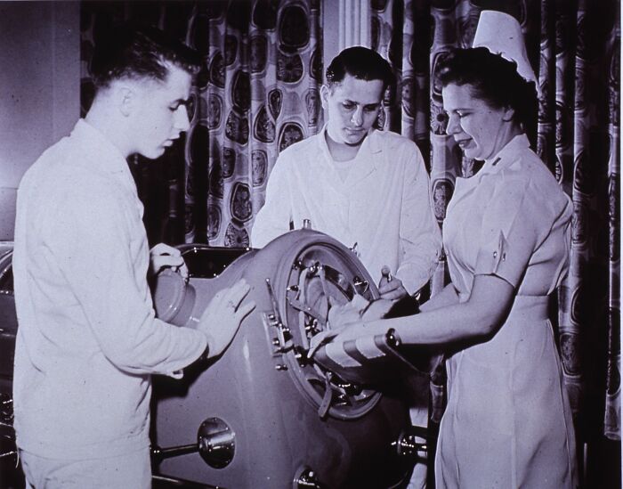 Three medical professionals using an old-school medical device to treat a patient’s injured arm in a vintage hospital setting.