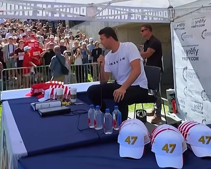 Man speaking into microphone at outdoor event with crowd behind, related to nurse suspended for calling out doctor remarks.