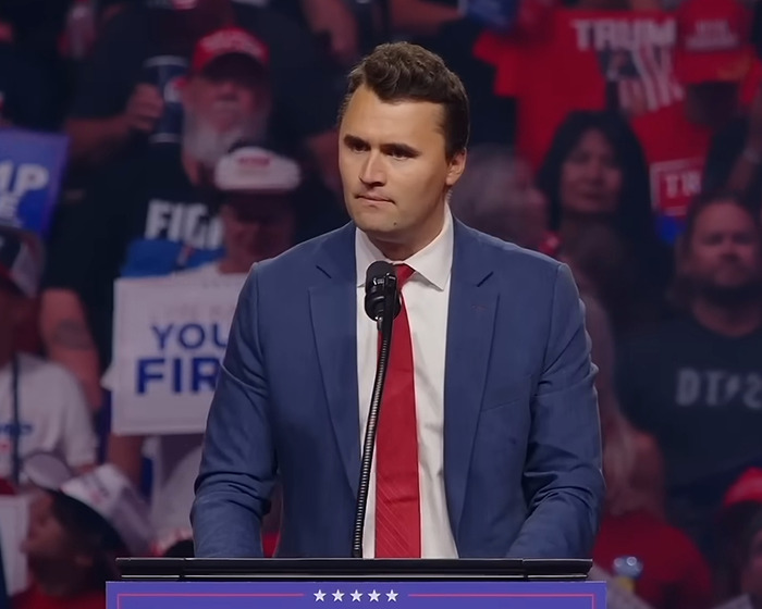 Charlie Kirk speaking at a podium in a blue suit and red tie during a crowded event with supporters in the background.