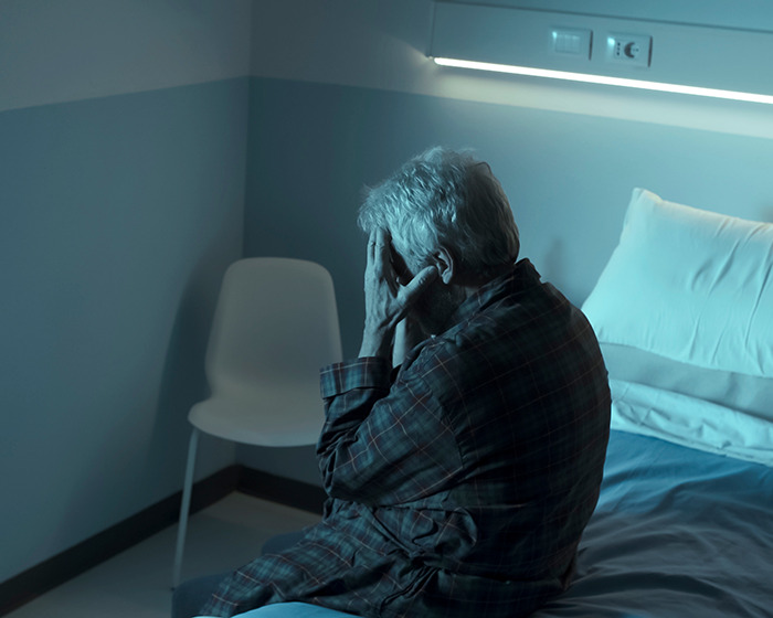 Elderly man sitting on a bed in a dim room, holding his head, representing the effects of dementia.